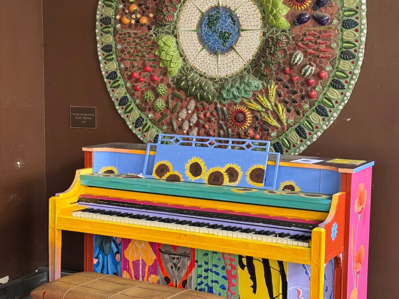 Colorfully painted piano with large, ceramic mandala on wall behind it at Davis Co op